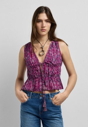 Jonge vrouw met lang bruin haar draagt een mouwloos paars topje met patroon en strikjes en een blauwe spijkerbroek, handen in de zakken, neutrale gezichtsuitdrukking.