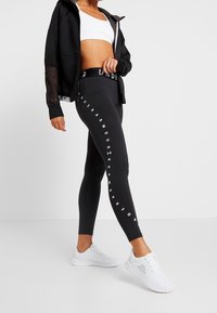 Black leggings with white branding along the side, paired with a white crop top and a black zip-up hoodie featuring mesh sleeves. White athletic shoes.