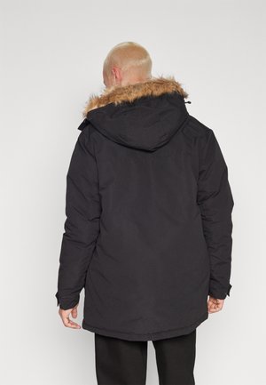 Mand med kort blondt hår iført en sort vinterfrakke med pelsforet hætte, set bagfra mod en ensfarvet baggrund.