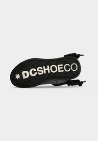 Semelle de chaussure noire avec un motif texturé montrant le texte blanc en relief DC SHOE CO et le logo étoile, lacets tressés noirs visibles sur les côtés.
