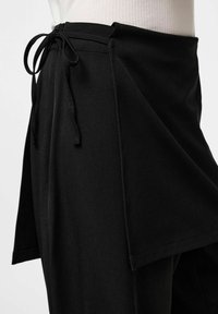 Jupe portefeuille noire avec une fermeture à nouer à la taille, confectionnée dans un tissu doux à la texture lisse et dotée d'un ourlet asymétrique.