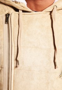 Giacca in suede beige con cappuccio foderato, zip frontale e cordini. Tessuto liscio con un leggero lucido e cuciture oblique.