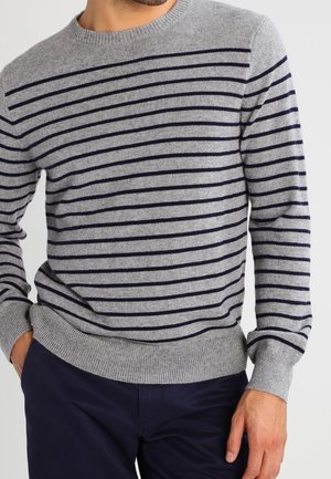 Uomo che indossa un maglione grigio girocollo con sottili strisce orizzontali scure e pantaloni blu scuro, in piedi con una mano in tasca.