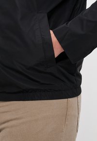 Giacca nera leggera con orlo elasticizzato, dotata di tasca laterale e texture liscia. Pantaloni marrone chiaro parzialmente visibili sotto.