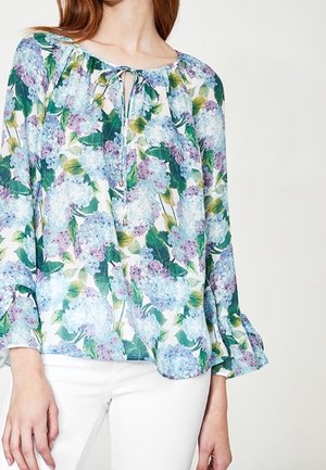 Femme vêtue d'un chemisier à manches longues avec imprimé floral bleu et violet et col à nouer, associé à un pantalon blanc sur fond uni.