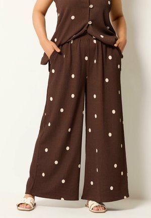 Femme portant un pantalon marron à jambes larges et un haut sans manches boutonné assorti à pois blancs, mains dans les poches, sandales blanches.