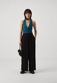 Teal halter top s hlbokým V-výstrihom, kombinovaný s čiernymi voľnými nohavicami. Doplnky zahŕňajú čiernu kabelku a čierny náhrdelník s detailom kvetu.