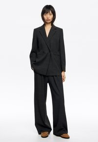 Blazer a cuadros gris oscuro con cierre de doble botonadura y dos bolsillos delanteros, combinado con pantalones sueltos a juego y zapatos marrones.