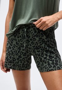 Personne portant un short à imprimé léopard vert avec un cordon de serrage et un haut olive ample, ajustant la taille avec la main.
