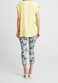 Ljusgul, kortärmad topp med avslappnad passform, kombinerad med blå byxor i slim-fit med blommönster och vita klackskor med blockhäll.