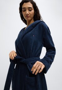 Μπλε ναυτικό μπουρνούζι κατασκευασμένο από υφασμα με υφή, με κουκούλα, δεμένο στη μέση. Διαθέτει δύο μπροστινές τσέπες και μια μικρή λεπτομέρεια με λογότυπο στο στήθος.