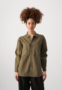 Olive green button-up shirt with long sleeves, featuring a chest pocket, textured fabric, and a relaxed fit. Paired with black striped pants.