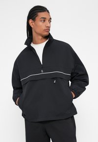 Giacca nera con mezza zip e colletto alto, polsini elastici, due tasche frontali e una striscia orizzontale bianca sul petto. Materiale leggero.