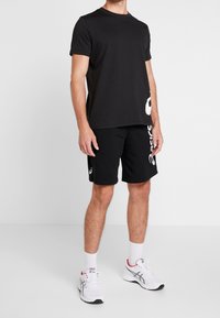 Black short-sleeve t-shirt with a crew neck, paired with black shorts featuring a white logo print. Worn with white athletic shoes and socks.