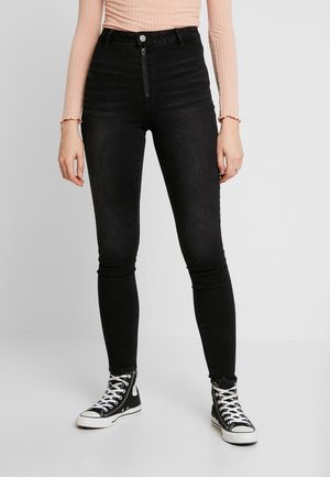 Kobieta nosząca czarne obcisłe dżinsy z wysokim stanem i suwakiem, jasnoróżową bluzkę z długim rękawem oraz czarne wysokie trampki z białymi sznurówkami.