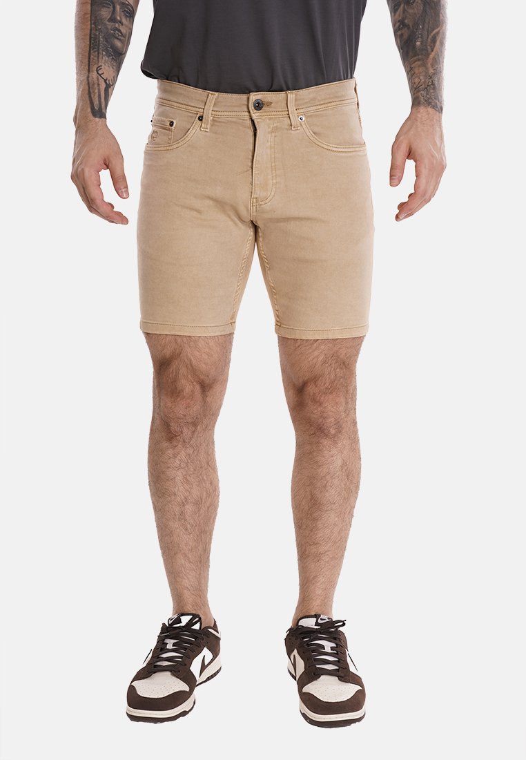 Shorts in denim color sabbia con un design aderente, cuciture sottili, cinque tasche e hardware metallico. Indossati con scarpe da ginnastica in bianco e nero.