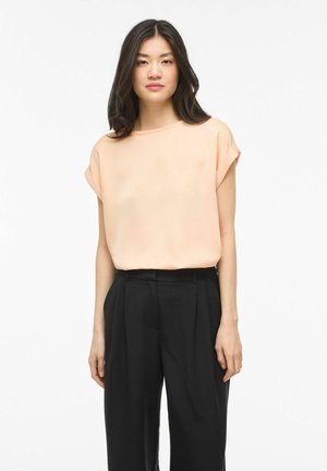 Ung kvinne med langt mørkt hår iført en løs ferskenfarget kortermet topp og høytlivs svarte bukser, stående mot en hvit bakgrunn.