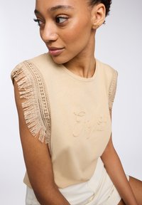 T-shirt beige con dettagli a frange sulle maniche e testo ricamato "Enjoy". Tessuto morbido con una vestibilità comoda e collo tondo.