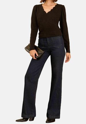 Kvinde iført sort top med lange ærmer og bølget halsudskæring, mørkeblå højtaljede vidde jeans, sorte hælede sko, holder en sort clutch.