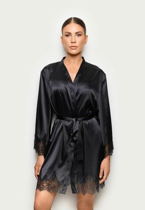 Donna che indossa un accappatoio di raso nero con rifiniture in pizzo su maniche e orlo, legato in vita, in piedi davanti a uno sfondo chiaro e uniforme.