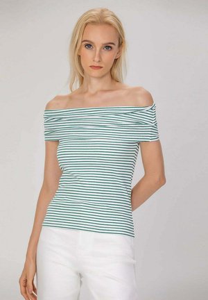 Femme blonde portant un haut blanc épaules dénudées avec des rayures vertes et un pantalon blanc, debout devant un fond clair uni.