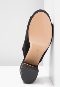 Chaussure à talon haut en daim noir inclinée vers le haut, montrant une semelle lisse beige clair avec le numéro de taille 4 et un appui talon texturé.