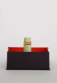 Mörk rektangulär låda med röd inredning som håller en flaska med grön lock märkt "pixi Glow Tonic." Flaskan är transparent med en orange vätska.