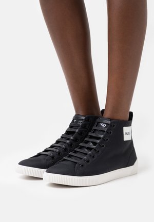 Schwarze High-Top-Sneaker mit weißen Sohlen und Schnürsenkeln, getragen an Beinen mit dunklem Hautton, mit einem weißen "HUGO" Logo-Patch an der Seite.