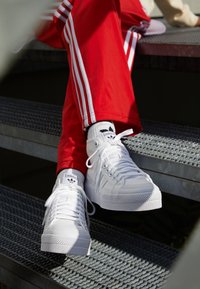 Zapatillas de lona blancas con suelas texturizadas, que cuentan con cordones prominentes y un logo de Adidas en la lengüeta, combinadas con pantalones de chándal rojos.