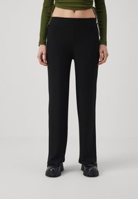 Vrouw in hoogzittende zwarte geribbelde broek, zwarte gestifte laarzen en een cropped langemouwen olijfgroene top, staande voor een effen achtergrond.