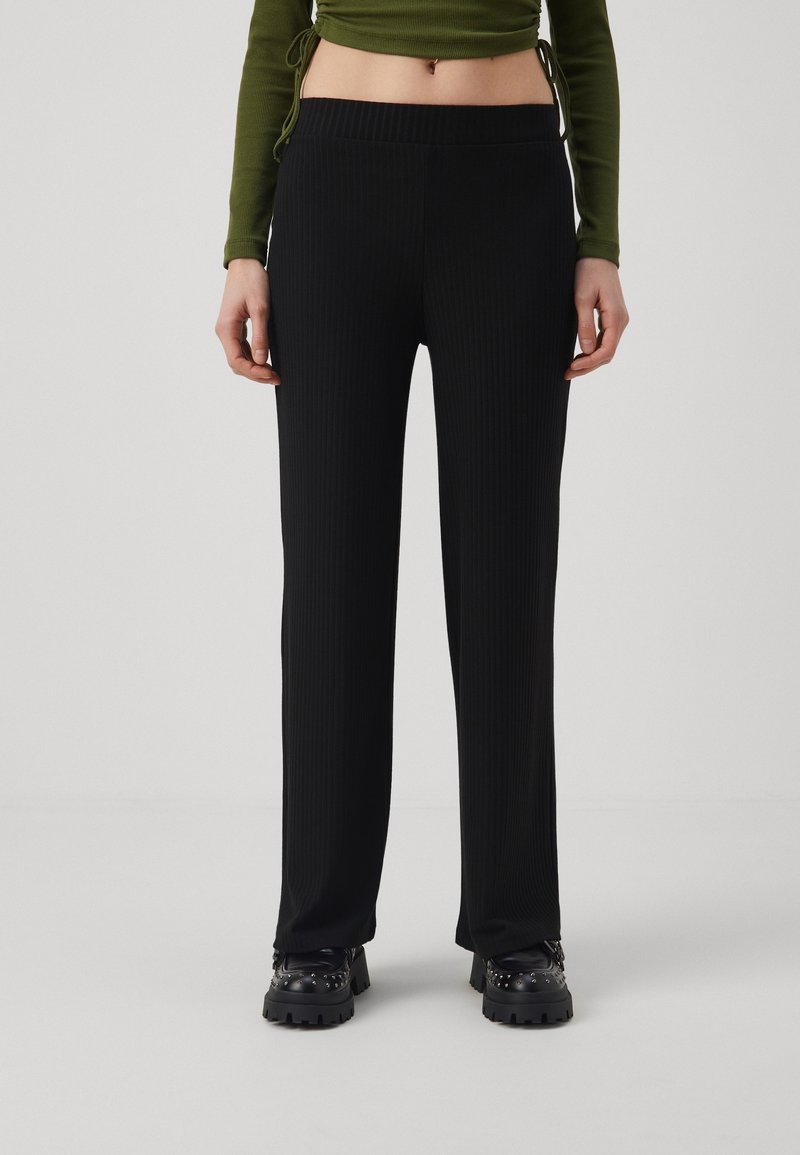 Vrouw in hoogzittende zwarte geribbelde broek, zwarte gestifte laarzen en een cropped langemouwen olijfgroene top, staande voor een effen achtergrond.
