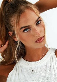 Top bianco lavorato a maglia con un motivo texture, abbinato a una collana con pendente argentato. Capelli biondi lunghi e mossi parzialmente raccolti. Occhi azzurri.