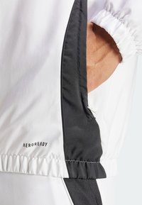 Biała kurtka z czarnymi panelami bocznymi, z nadrukowanym logo na dolnym brzegu. Materiał wydaje się lekki; konstrukcja zawiera elastyczne mankiety i pas.