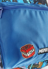 Funda de tela azul con una superficie texturada, con una etiqueta de TNT roja y la palabra "MINE" en negro. Cierre de cremallera con detalles en contraste.