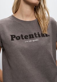 Grå bomullströja med strukturerad svart text som läser "Potential." med mindre vit text under, "du är kapabel."