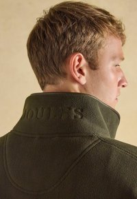 Tmavozelená fleecová bunda s vysokým golierom. Má embosované písmo na chrbte, hladkú textúru a jemné švy.