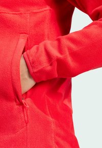 Nahaufnahme einer roten Fleecejacke, die eine Hand zeigt, die in eine Seiten Tasche greift. Die Jacke hat eine glatte Textur und elastische Bündchen.