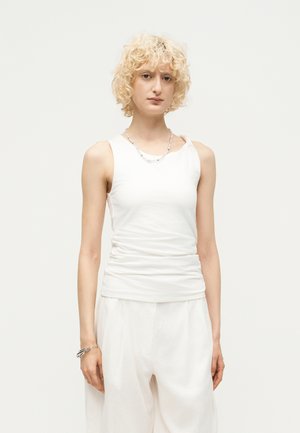 Jeune personne aux cheveux blonds courts et bouclés, portant un haut blanc sans manches et un pantalon blanc large, debout devant un fond clair uni.