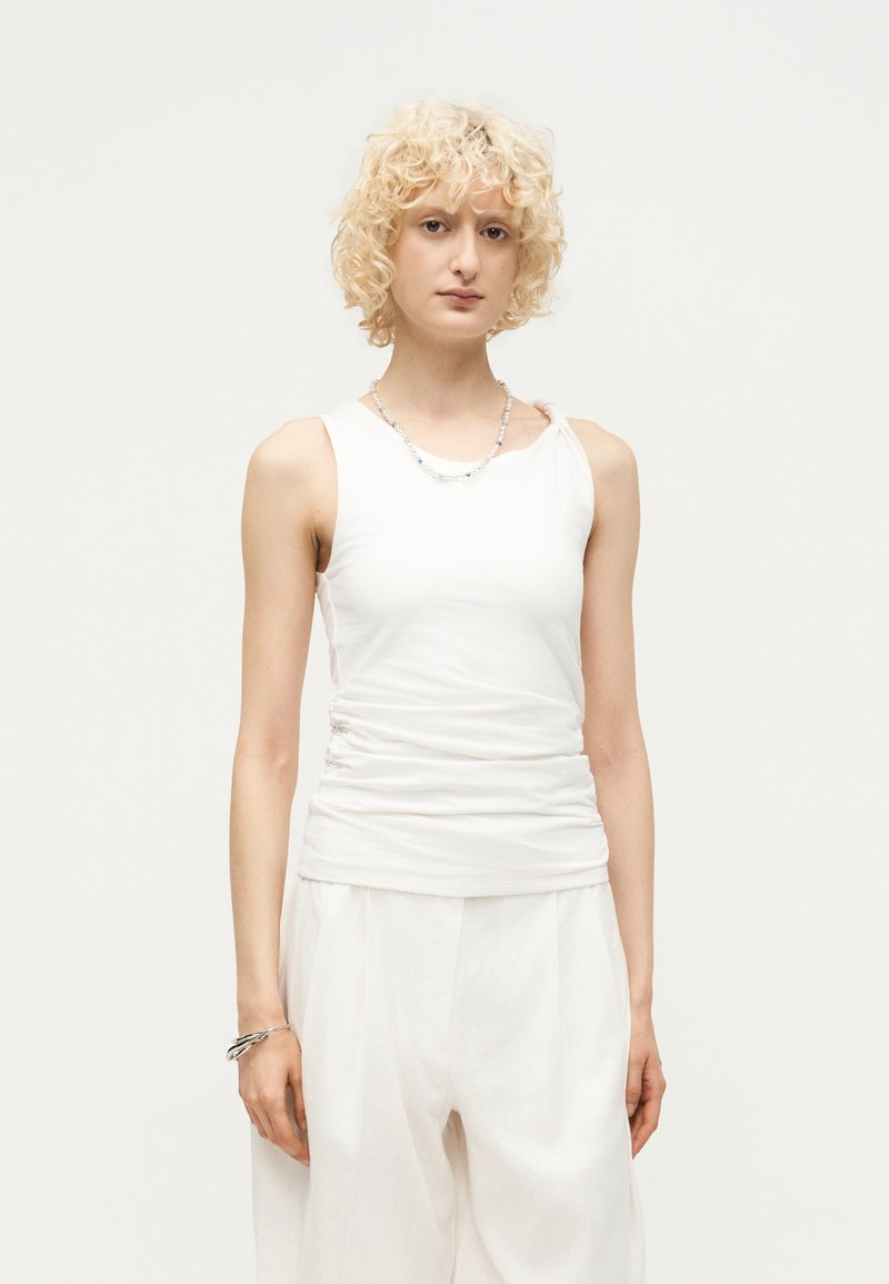 Νέο άτομο με κοντά σγουρά ξανθά μαλλιά που φοράει αμάνικο λευκό τοπ και φαρδιά λευκά παντελόνια, στέκεται μπροστά σε ένα απλό φωτεινό φόντο.