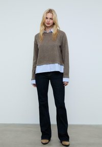 Brauner Wollpullover, über einem hellblau gestreiften Hemd getragen. Dunkelblaue, ausgestellte Jeans und hellbraune Schuhe. Glatte Texturen und lässige Passform.