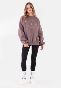 Oversize lawendowy sweatshirt z okrągłym haftowanym detalem i literą "C" na środku, w połączeniu z czarnymi legginsami i białymi trampkami.