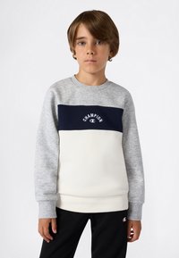 Chłopiec z jasnobrązowymi włosami, ubrany w szary, granatowy i biały sweatshirt Champion oraz czarne spodnie na jednolitym białym tle.