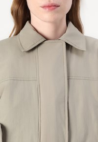 Svijetlosiva, teksturirana jakna s širokim ovratnikom, vertikalnim šavovima i zatvaranjem na zip. Tkanina izgleda glatko i lagano.