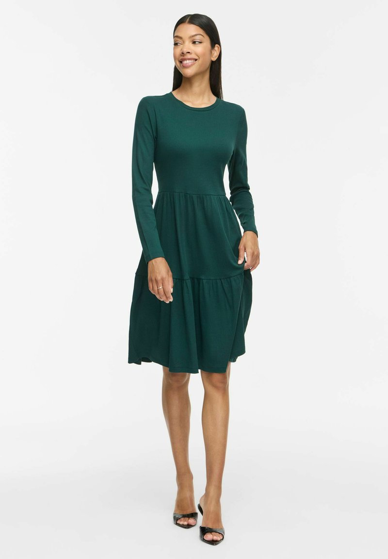 Femme debout et souriante, vêtue d'une robe verte foncée à manches longues et longueur genou, avec des talons ouverts noirs sur fond blanc.