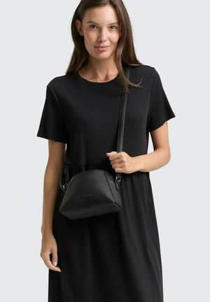Schwarze Leder Schultertasche mit strukturiertem Finish, geschwungenem Design, silberner Hardware und abnehmbarem Tragegurt, gehalten von einer Person in einem schwarzen Kleid.