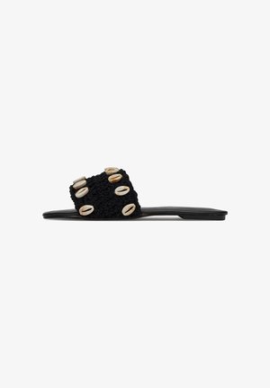 Schwarze Rutsch-Sandale mit einem dicken gewobenen Stoffriemen, der mit fünf Kaurischalen verziert ist, auf einer flachen schwarzen Sohle.
