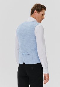 Kamizelka w jasnoniebieski wzór houndstooth, materiał o subtelnej teksturze, guziki z tyłu, połączona z białą koszulą z długim rękawem i czarnymi spodniami.