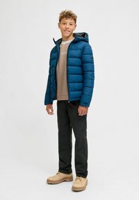 Un adolescent se tenant debout, portant une veste matelassée bleue avec capuche, un sweat-shirt beige, un pantalon cargo noir et des bottes lacées couleur sable.