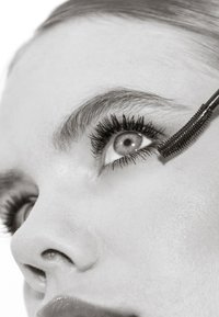 Ingrandimento di una persona che applica il mascara sulle ciglia superiori con un scovolino curvo, mettendo in risalto i dettagli del trucco per gli occhi.
