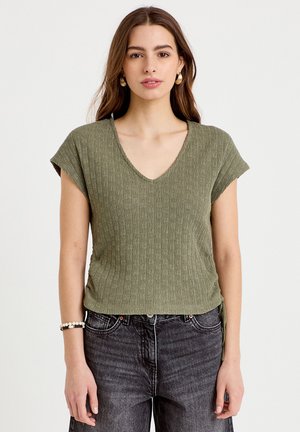 Jonge vrouw die een olijfgroene gebreide top met korte mouwen en V-hals draagt, samen met donkergewassen jeans, staand voor een effen witte achtergrond.